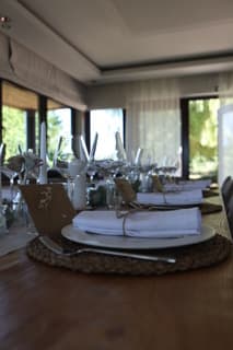 Longue table de bois dressée, serviettes blanches nouées de ficelle sur des sets en osier, baies vitrées en fond