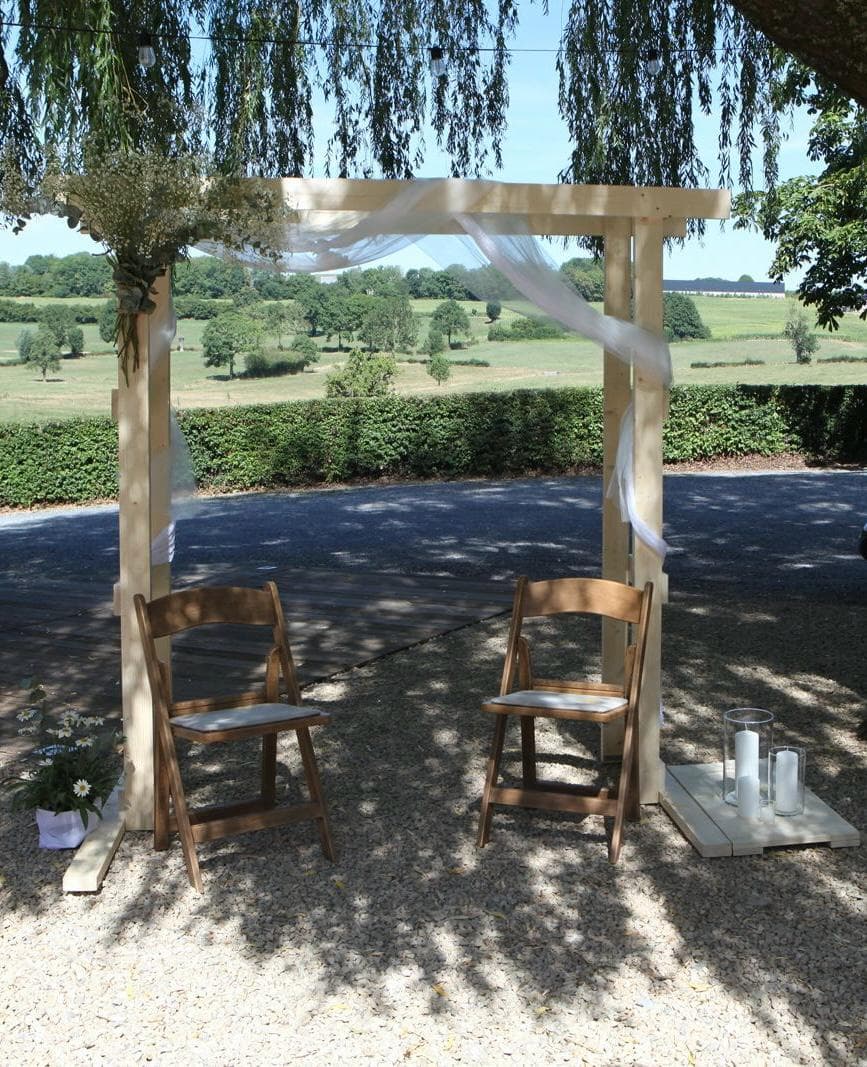 Arche de bois drapée de voile blanc, deux chaises pliantes et des photophores sur le gravier, la campagne en contrebas